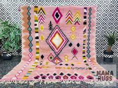 a pink rug with multicolored diamonds and fringes on the floor next to a potted plant