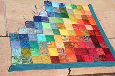 a multicolored quilt is laying on the ground next to a brick wall and sidewalk