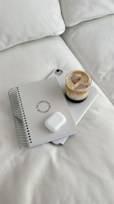 an open notebook, mouse and coffee cup sitting on a bed with white linens