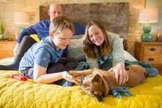 two people and a dog laying on a bed with yellow sheets, smiling at each other