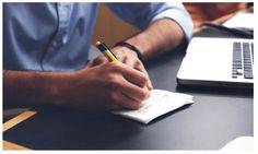 a man sitting at a desk writing on a notepad with a laptop in the background