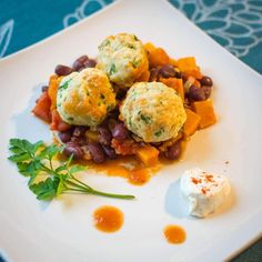 a white plate topped with meatballs and beans covered in sauce next to a small dollop of sour cream