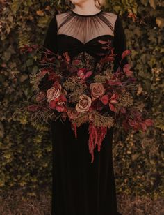 a woman wearing a black dress with red flowers on her chest and long sleeves, standing in front of a hedge