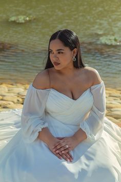 a woman in a white dress is sitting on the rocks by the water and posing for a photo