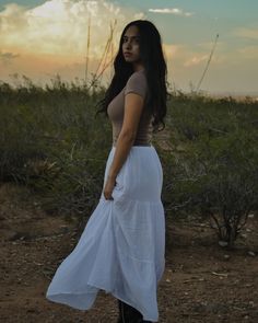 a woman is standing in the desert wearing a long white skirt and black cowboy boots