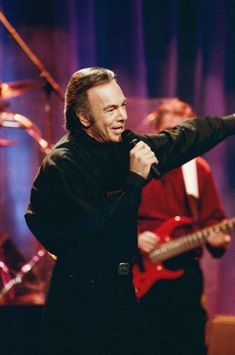 a man holding a microphone in front of two other men on stage with instruments behind him