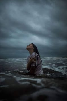 a woman sitting in the water with her eyes closed, looking up at an overcast sky