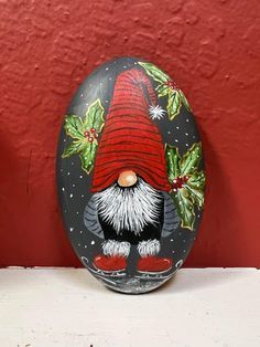 a painted rock with a gnome face and holly leaves on it, sitting against a red wall