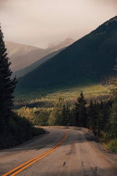 an empty road in the mountains with trees on both sides and snow capped mountains behind it