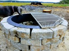 a dog standing next to a fire pit in the middle of a dirt field with a metal grate on it's side