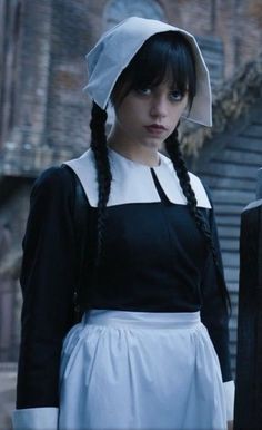 a woman with long black hair wearing a white bonnet and dress standing next to a brick building