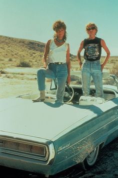 two people standing on top of a car in the desert