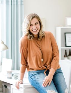 a smiling woman sitting on top of a white desk in front of a blue curtain