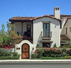 a white house with red flowers in the front yard and a brown door on the side