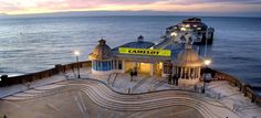 a pier that is next to the ocean at night with people walking around it and lights on