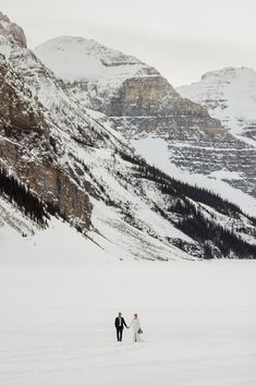 a bride and groom walk through the snow in front of some snowy mountains on their wedding day