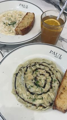 two white plates topped with pasta and bread next to a glass of orange juice on a marble table