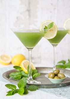 two martini glasses filled with green liquid and lemons on a tray next to mint leaves