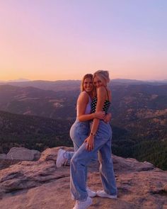 two women hugging each other while standing on top of a mountain at sunset with mountains in the background