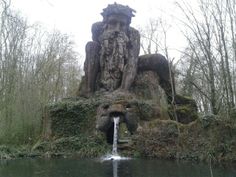 a fountain in the middle of a pond surrounded by trees and bushes with statues on it