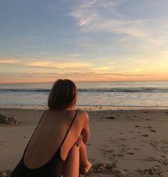 a woman sitting on top of a sandy beach next to the ocean in front of a sunset