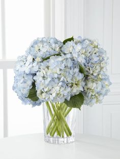 a glass vase filled with blue flowers on top of a white table next to a window