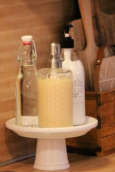 soap and lotion bottles are on a white cake plate next to other items in wooden boxes