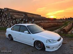 a white car parked in front of a pile of logs with the sun setting behind it