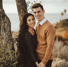 a man and woman standing next to each other under a tree with the ocean in the background