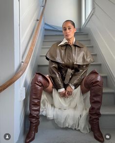 a woman sitting on the stairs wearing boots and a leather jacket with long sleeves over her shoulders