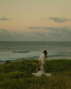 a man and woman standing on top of a lush green hillside next to the ocean