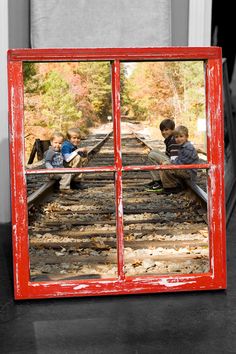 three children are sitting on the railroad tracks looking through a window at autumn foliage and trees