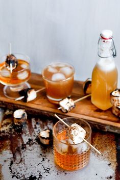 an assortment of drinks are sitting on a wooden tray with ice and marshmallows