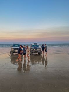 several people are standing in the sand next to two trucks on the beach at sunset