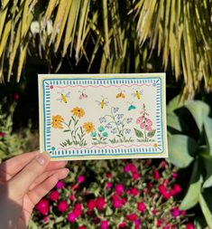 a person holding up a card with flowers and butterflies on it in front of some plants