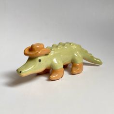 a ceramic toy alligator with an orange hat on its head and legs, sitting on a white surface