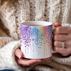 a woman is holding a coffee mug with watercolor paint on it and has her hands in the cup