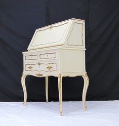 an antique white desk with gold trimmings on the top and bottom drawers, in front of a black backdrop