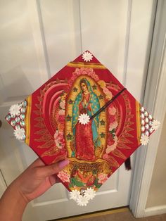 a hand holding up a red and gold graduation cap with an image of the virgin mary on it