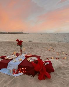 a table set up on the beach with candles and flowers in front of it at sunset