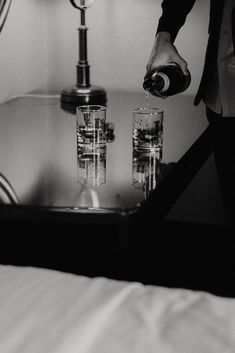 a person pouring water into two glasses in front of a lamp on a bed with white sheets