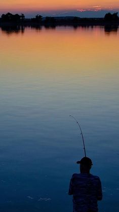 a man standing in the water while holding a fishing rod with a sunset behind him