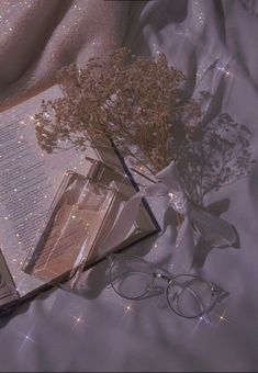 an open book on a bed next to some glasses and a plant with flowers in it