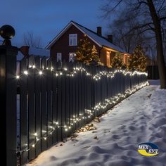 a fence with christmas lights on it in front of a house and trees covered in snow