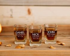 three shot glasses with personalized labels sit on a wooden table next to an orange