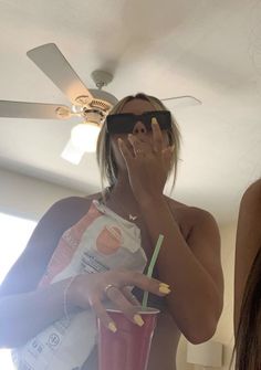 a woman wearing sunglasses holding a drink and talking on the phone while standing in front of a ceiling fan