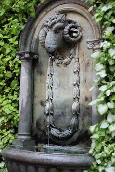 a stone fountain surrounded by greenery with a ram head on it's side