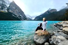 a woman sitting on top of a rock next to a body of water with mountains in the background