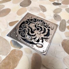 a square drain cover with water droplets on it and pebbles around it, sitting on top of a tiled floor