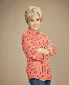 a woman with her arms crossed standing in front of a gray background wearing a pink shirt and jeans